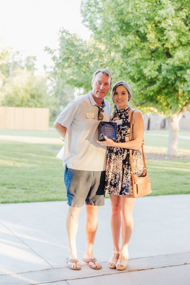 Man and woman standing outdoors, holding a plaque. Woman in floral dress, man in shorts, both smiling.