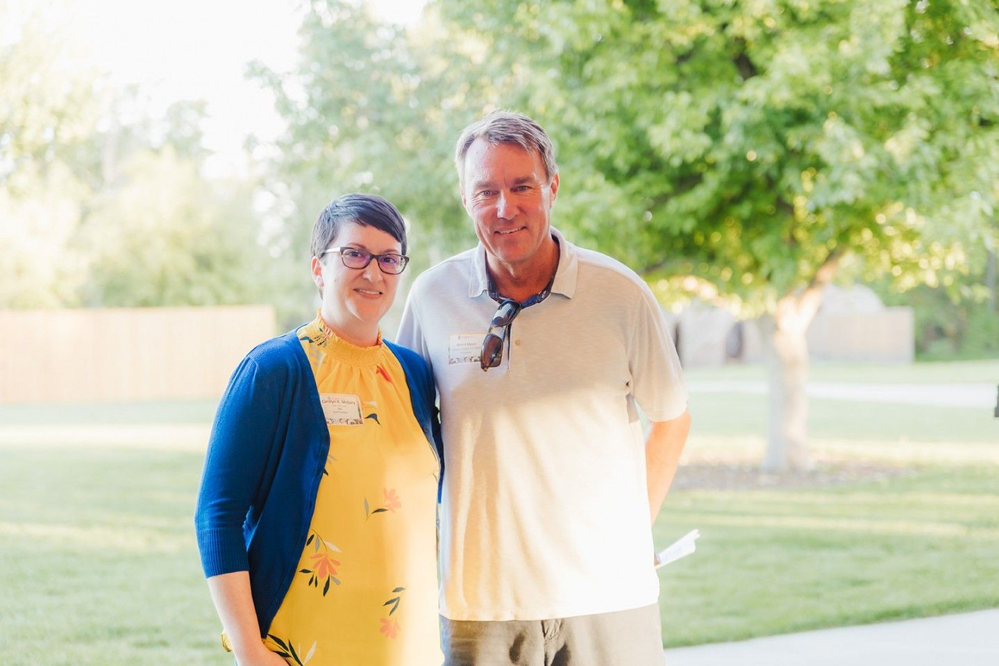 Woman in yellow dress and blue sweater poses next to a man in a polo shirt, outside, smiling.