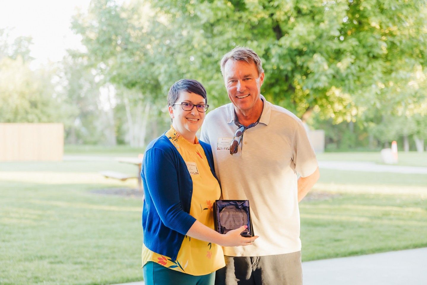 Woman in blue cardigan and man in polo shirt holding a plaque outdoors.