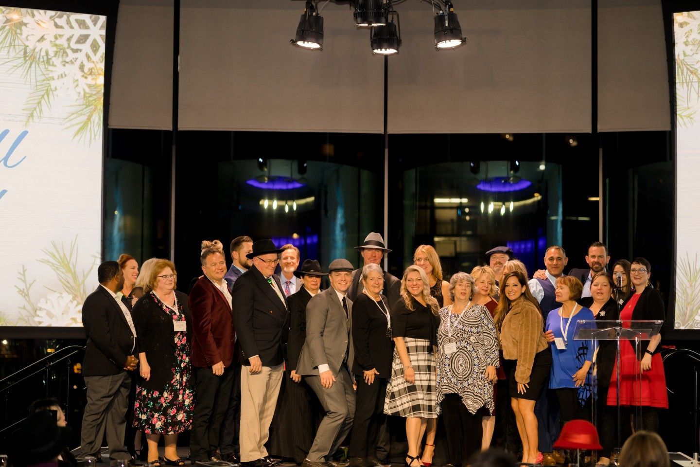 Group of people on stage, some in hats, at an event with lights and a backdrop.