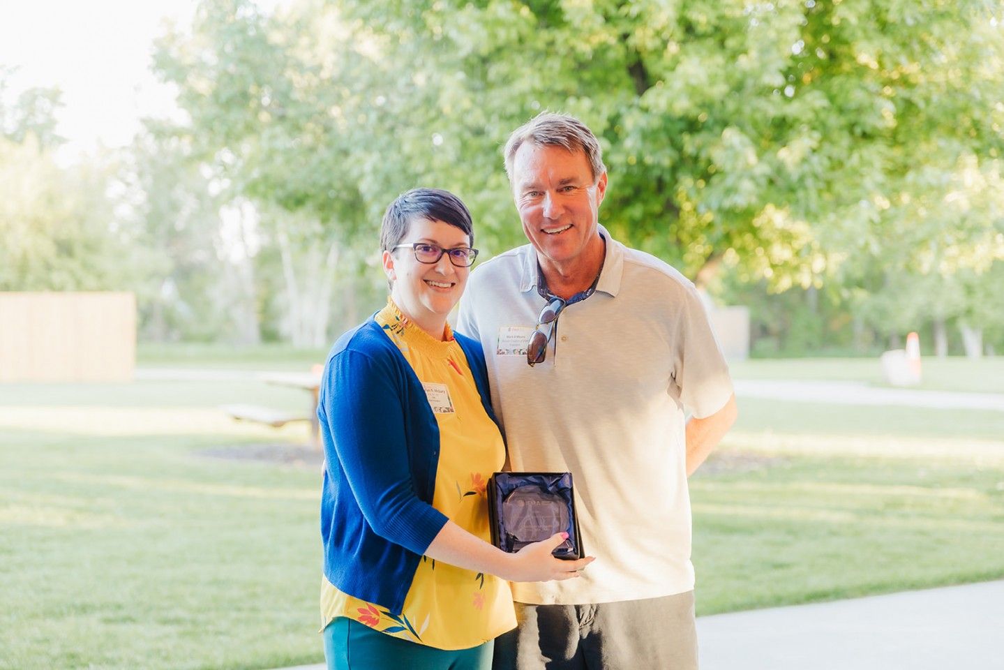 Woman in glasses presents an award to a man outdoors. Both smile.