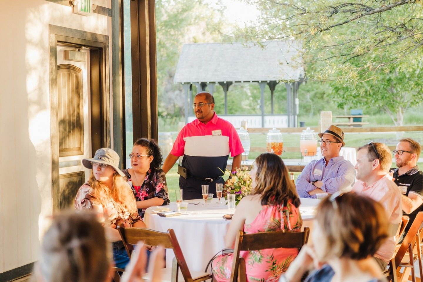 Man giving a speech to a seated group at an outdoor event, some people wear hats, white tablecloths, pavilion in background.
