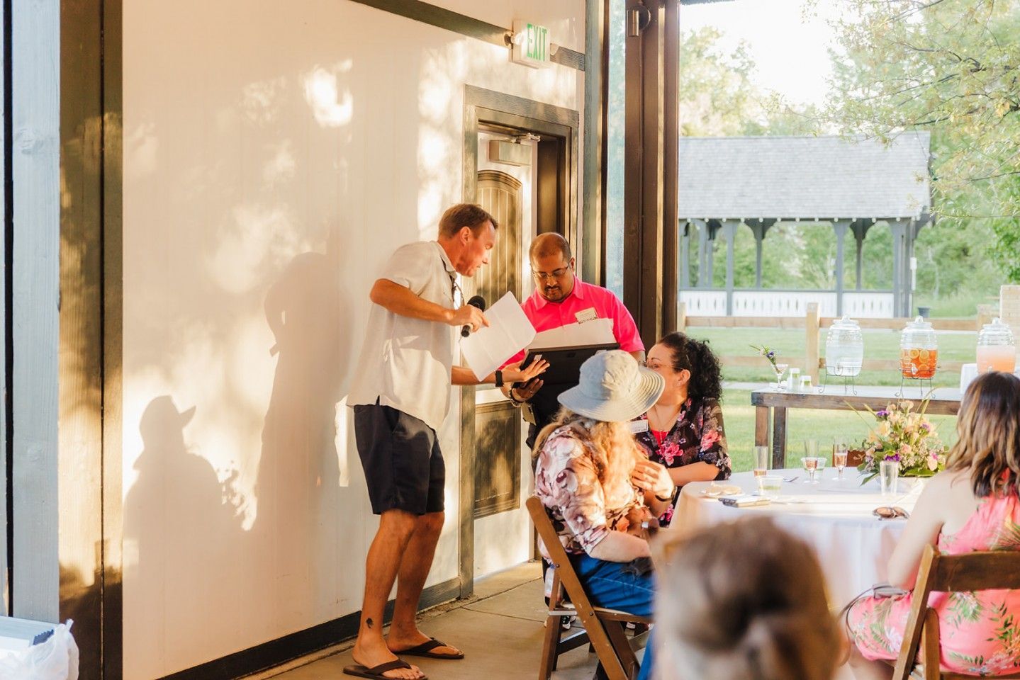 A man gestures while holding papers near an indoor event with others seated and a gazebo visible outside.