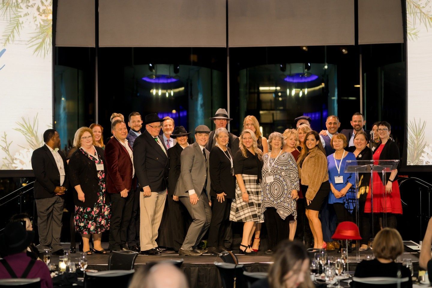 Group of people posing on a stage, some wearing hats. Backlit window, event setting.