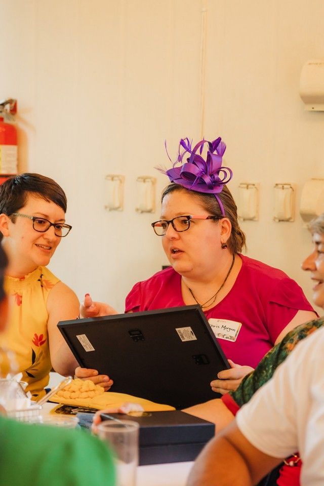 Woman in purple hat shows framed item to others at a table. Woman in yellow shirt smiles.