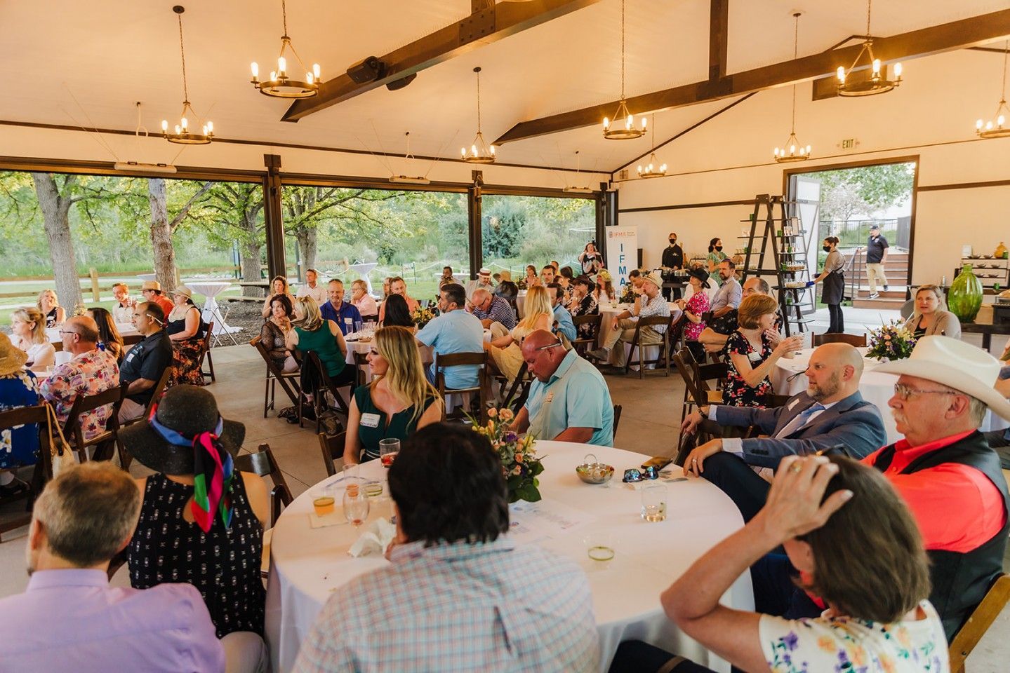 Guests seated at round tables under a pavilion, trees visible outside.