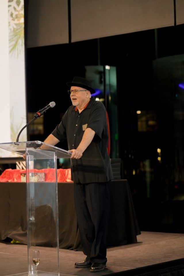 Man in black hat speaking at podium, night event.