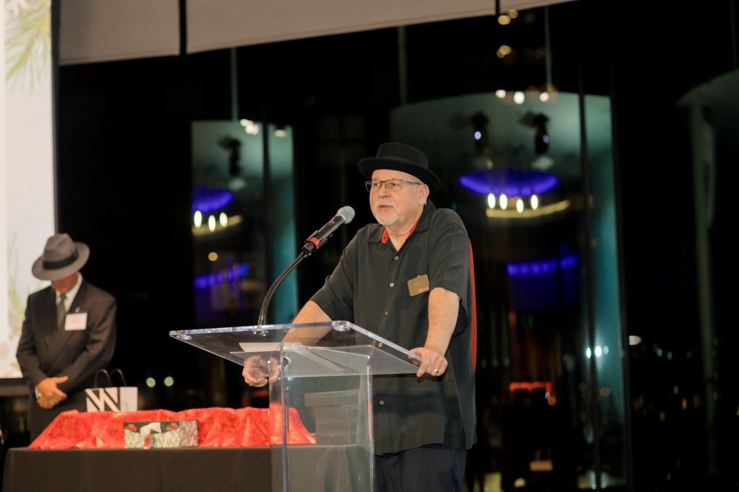Man in black hat speaking at a podium, another man in a suit stands by. Nighttime setting with city lights.