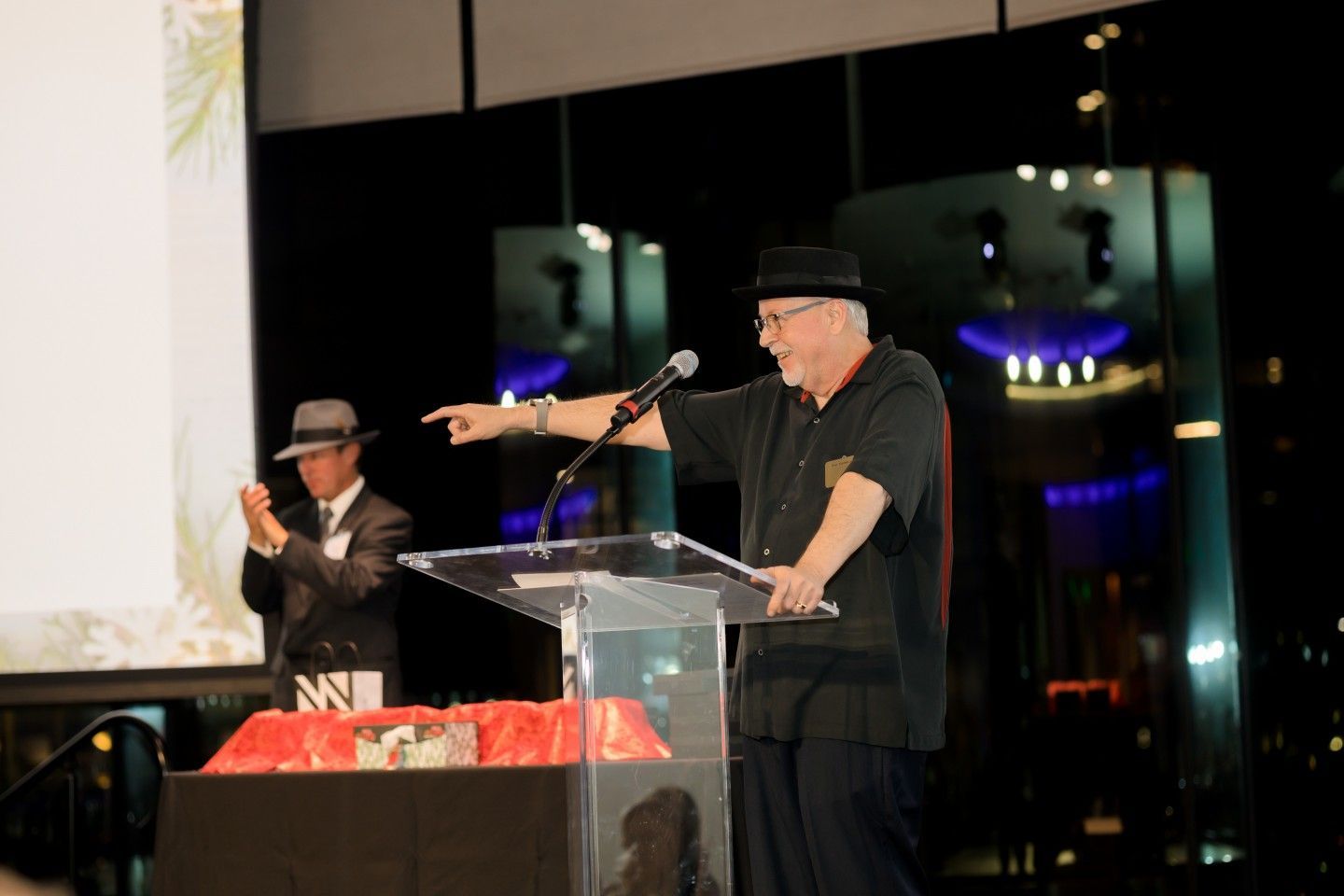 Man in black hat at podium gestures, addressing audience. Another man applauds behind him. Evening setting.
