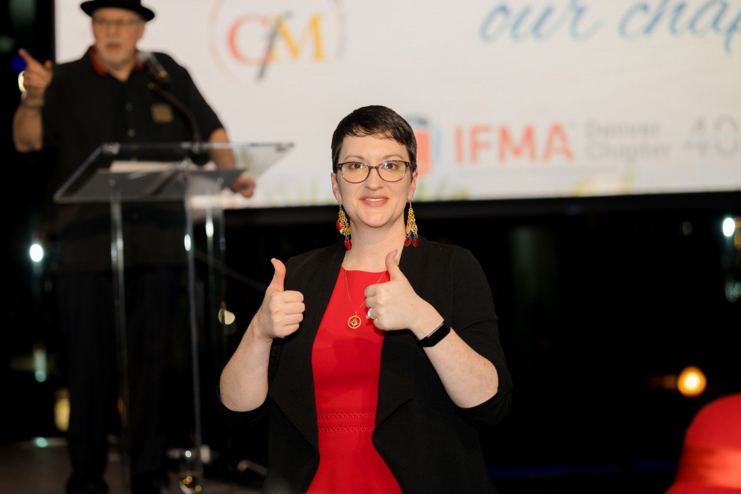 Woman in red dress and black blazer giving thumbs up at event with speaker.