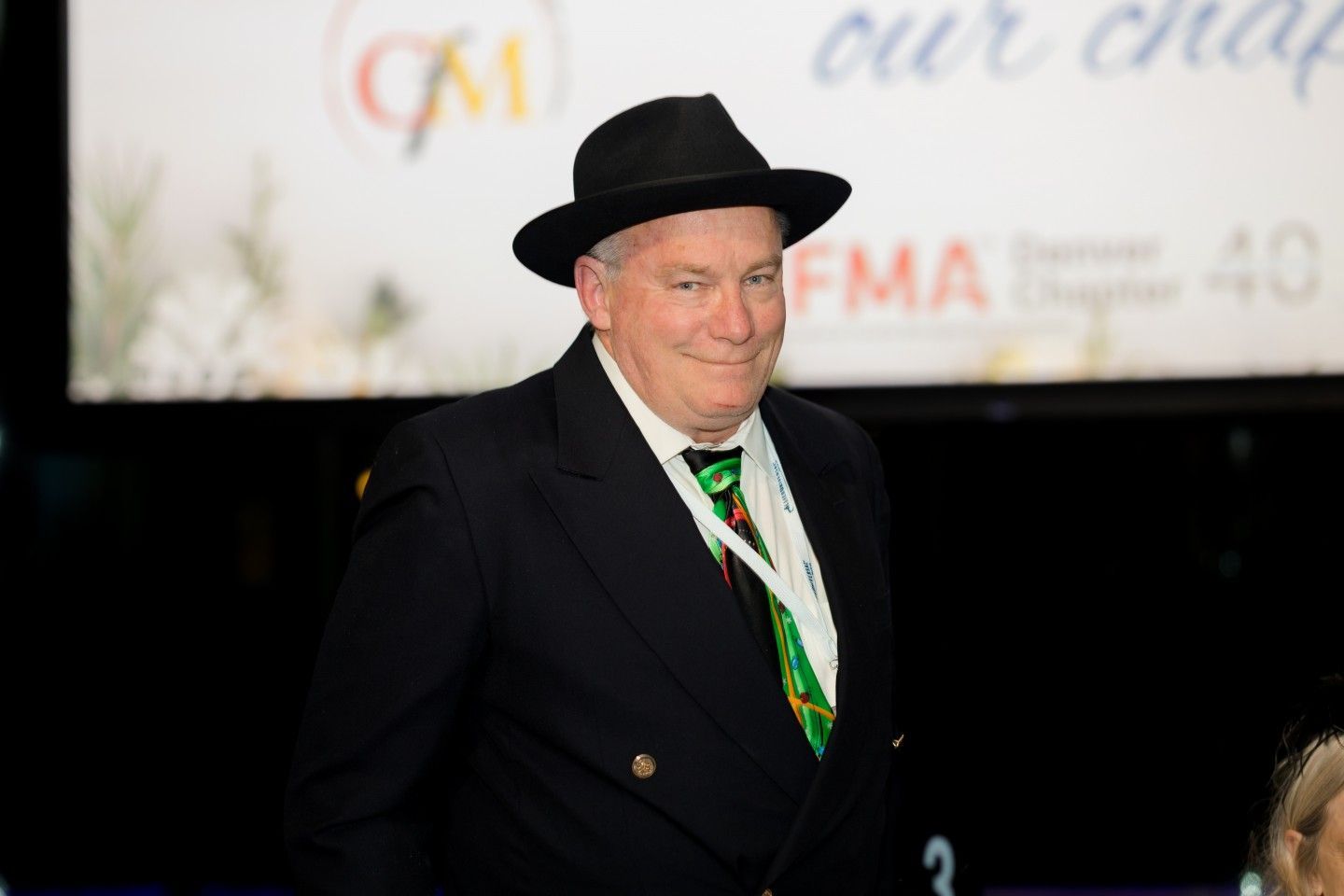 Man in black suit and hat, smiling at event, green tie and badge.