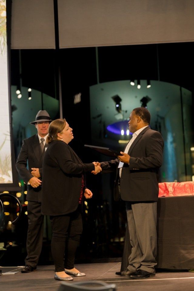 Woman receiving an award from a man onstage; a man in a hat watches.