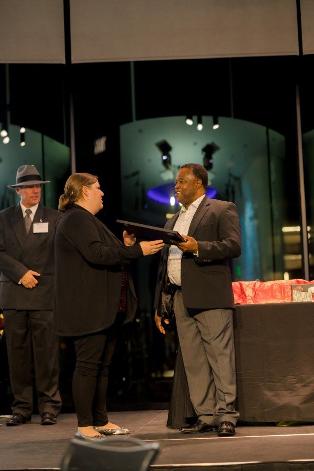 A person hands a document to another on a stage. A man in a suit and hat stands to the left.