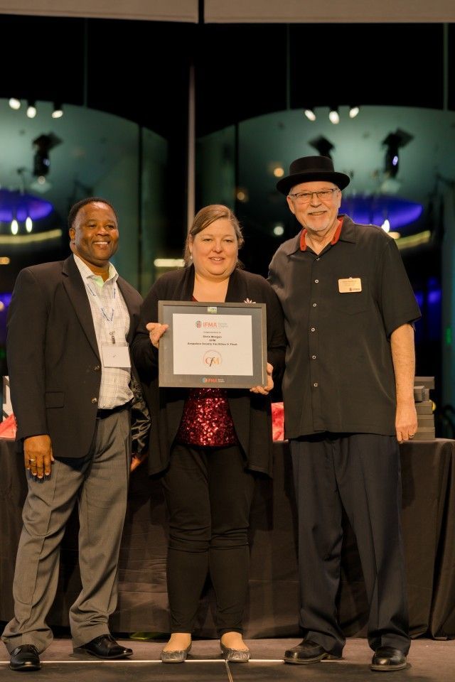 Three people on a stage holding an award. Woman in the center. Dark attire and stage background.