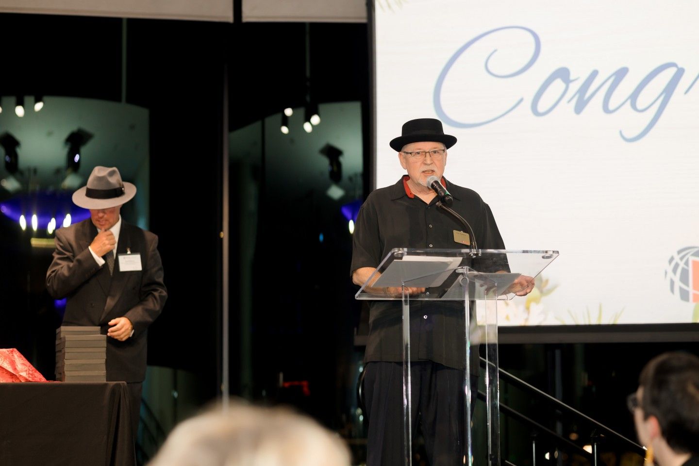 Man in hat speaking at a podium; another man stands nearby. 