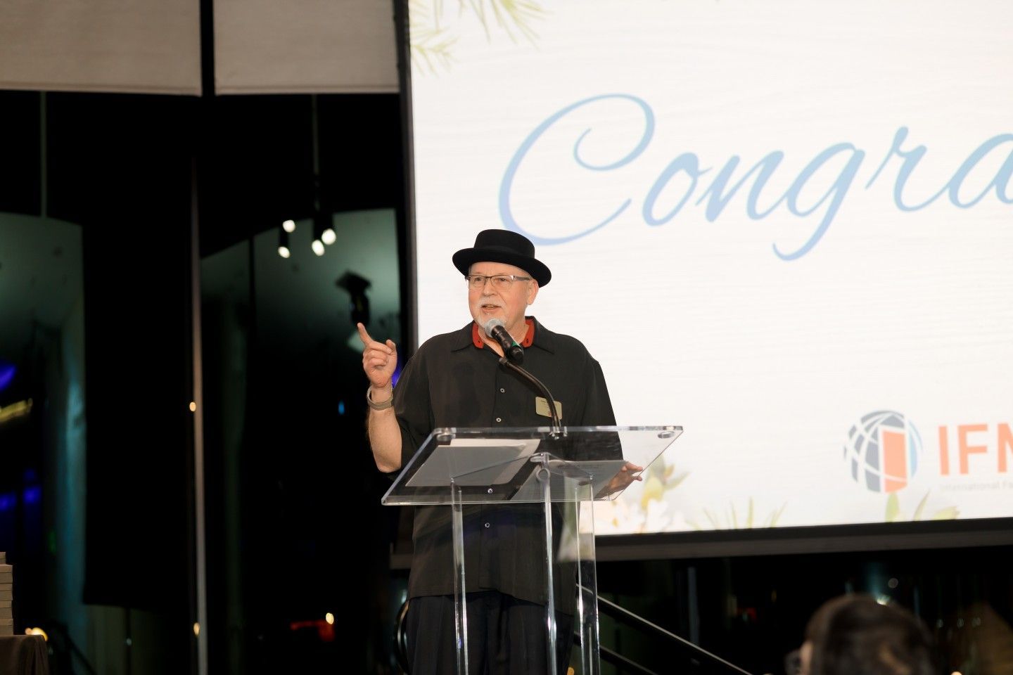 Man in black hat speaking at a podium with “Congratulations” on screen.
