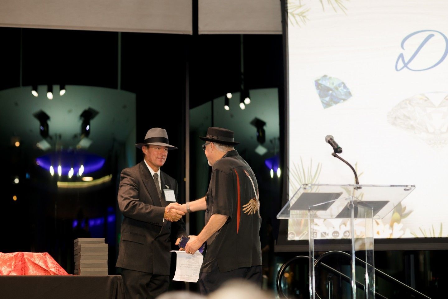 Two men in suits shake hands on a stage with a podium and a screen in the background.