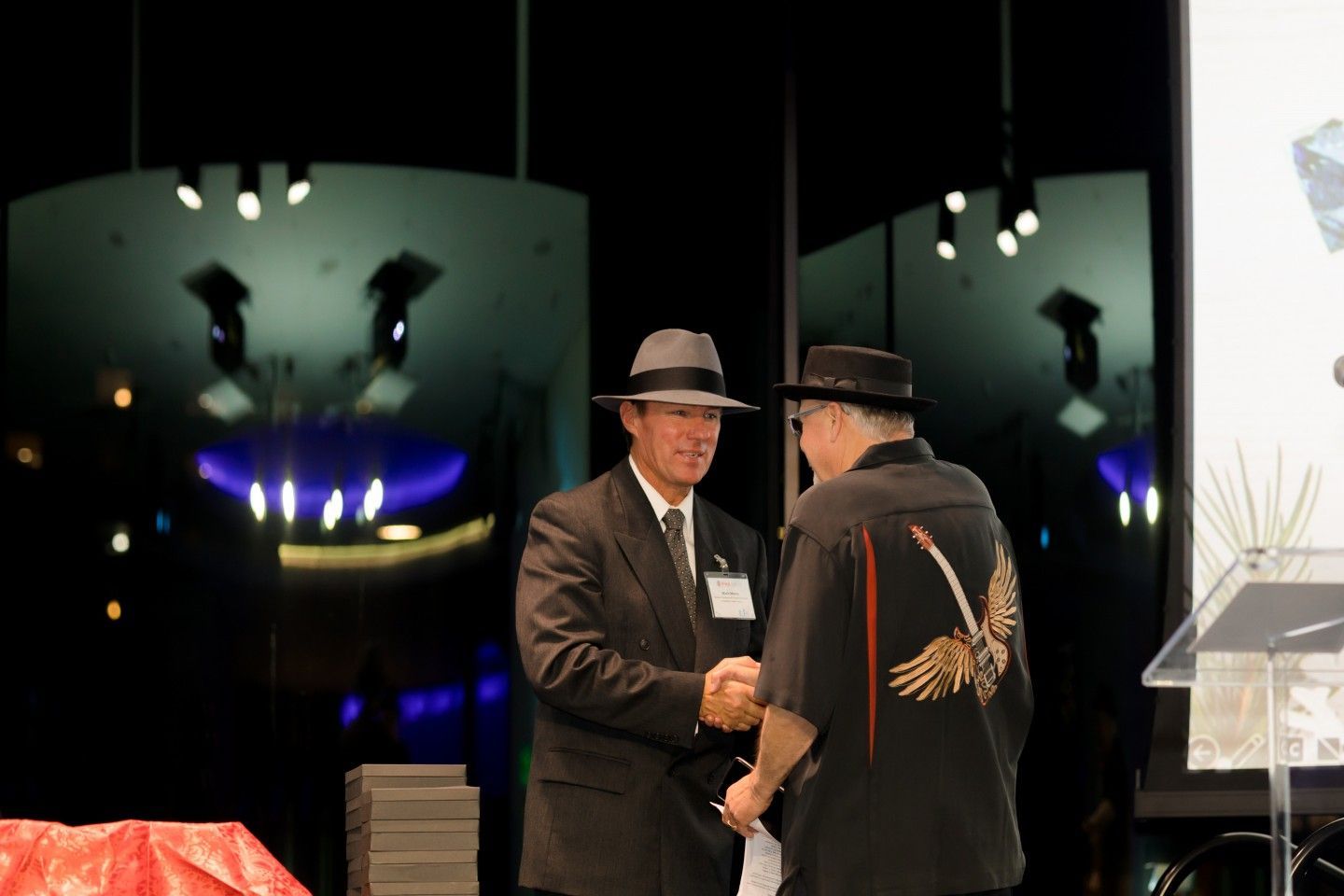 Two men shaking hands on stage; one in a suit, the other in a decorated shirt. Black background with stage lights.