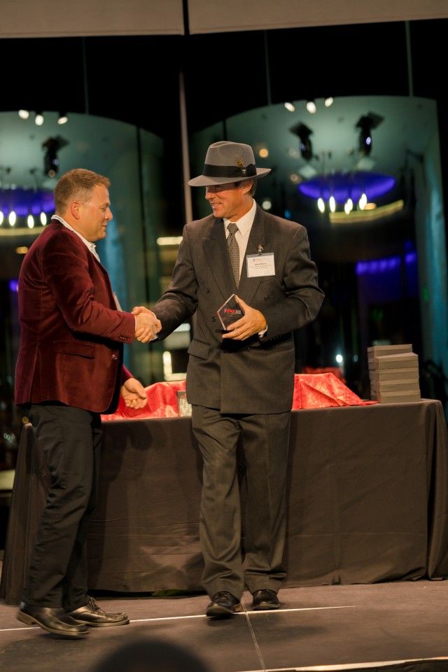 Man in burgundy jacket shakes hands with man in a suit and hat, receiving an award on stage.