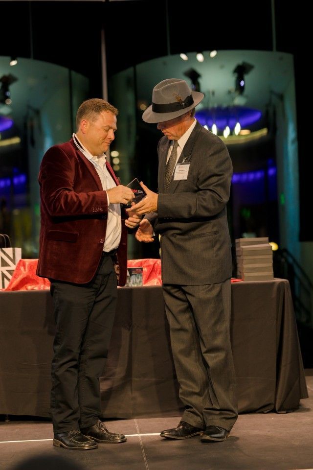 Man in maroon blazer presents award to man in suit and fedora on stage.