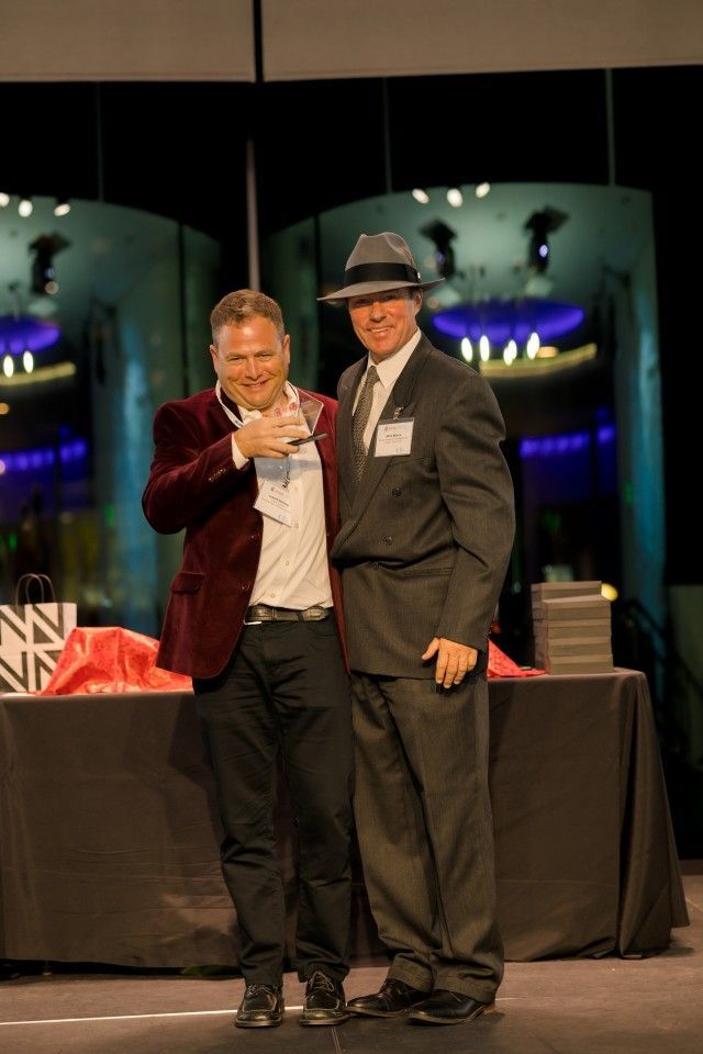 Two men on a stage; one in red jacket, holding award. The other in a suit, wearing a fedora.