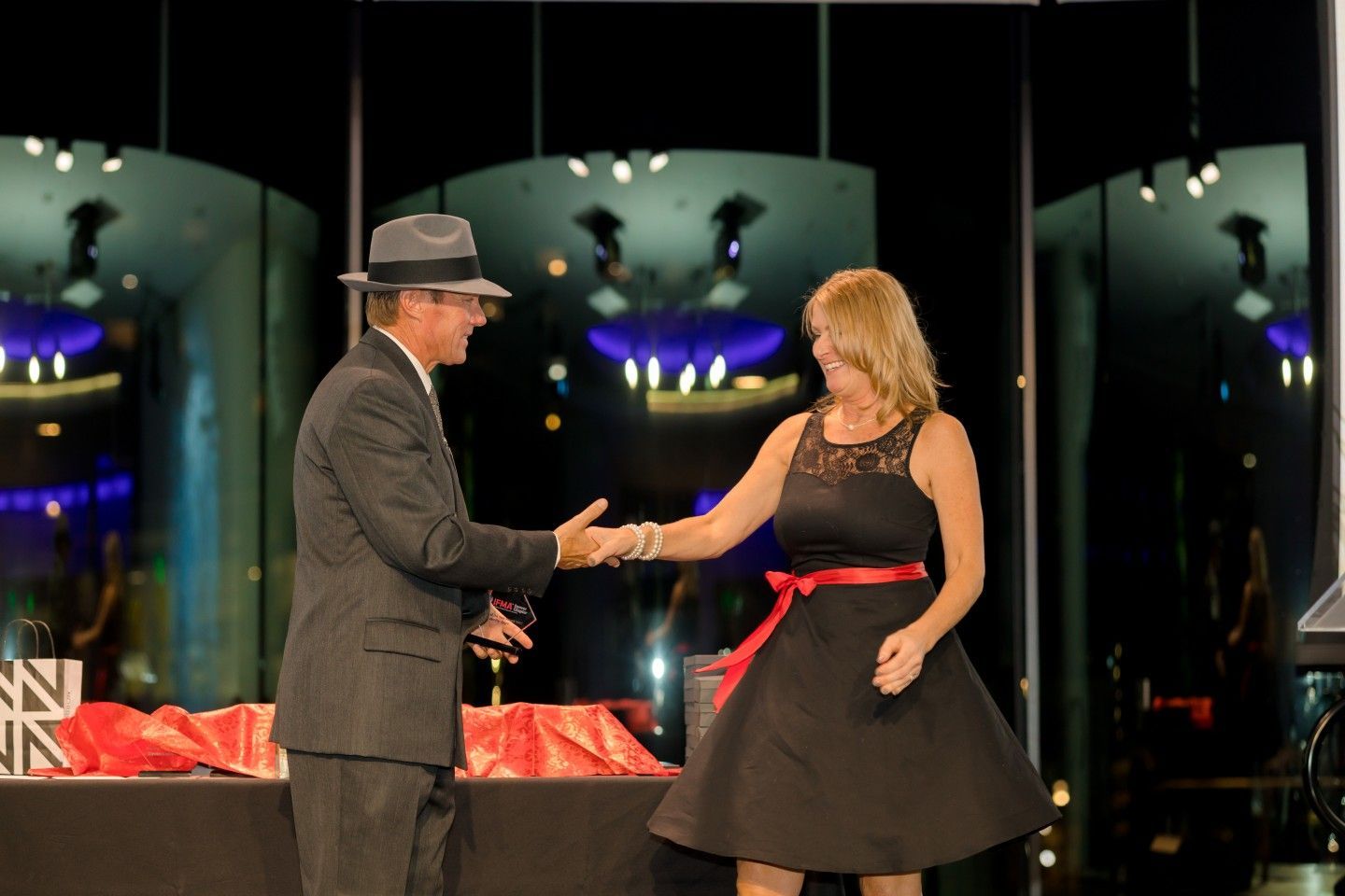 Man in hat and suit shaking hands with woman in black dress with red sash on stage.