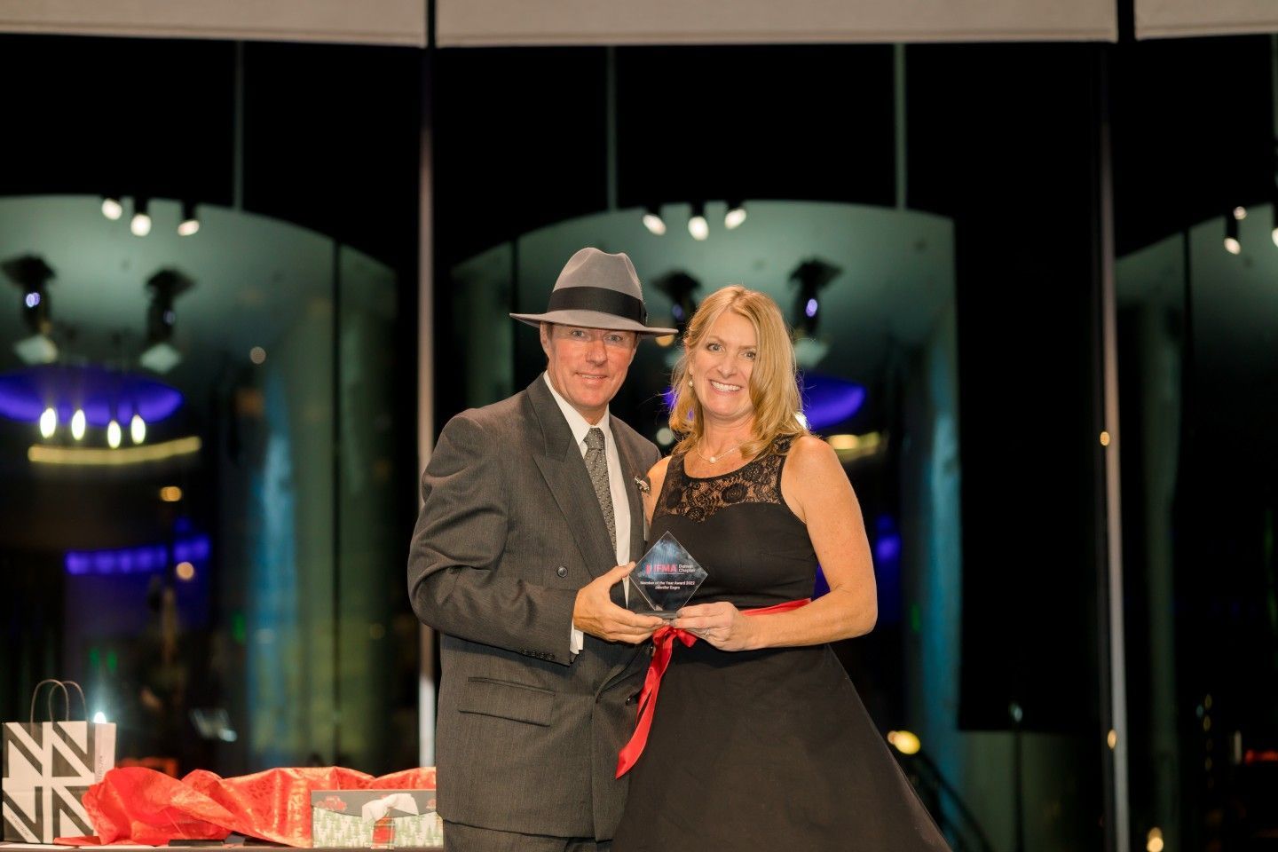 Man in suit and fedora presents award to woman in black dress, red sash. Interior with illuminated backdrop.