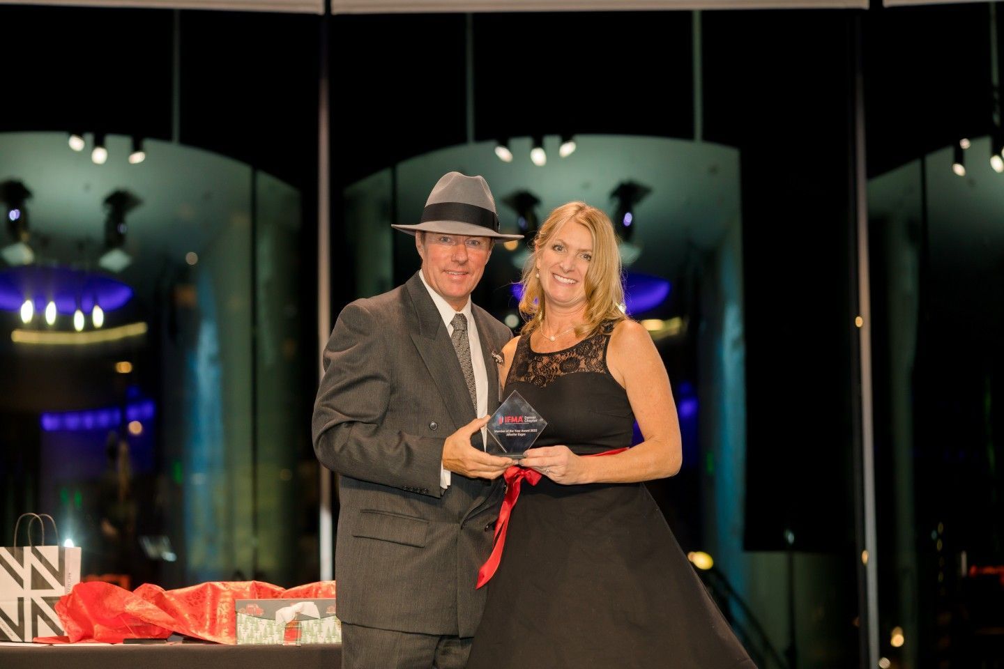 Man in suit and fedora presents award to woman in black dress.