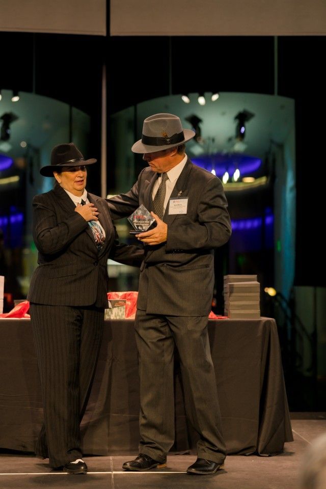 Two people in suits and hats; man holding award. Woman places hand on chest. Stage setting.
