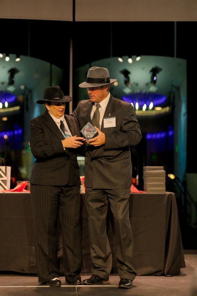 Two people in fedora hats and suits on stage, one holding an award.