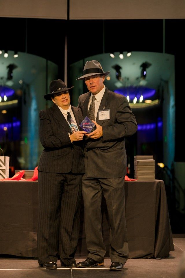 Two people in suits and fedora hats at an event, holding an award.