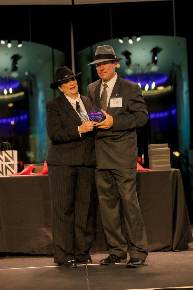 Two people in fedoras and suits on stage, holding an award, smiling. Stage backdrop with lights.