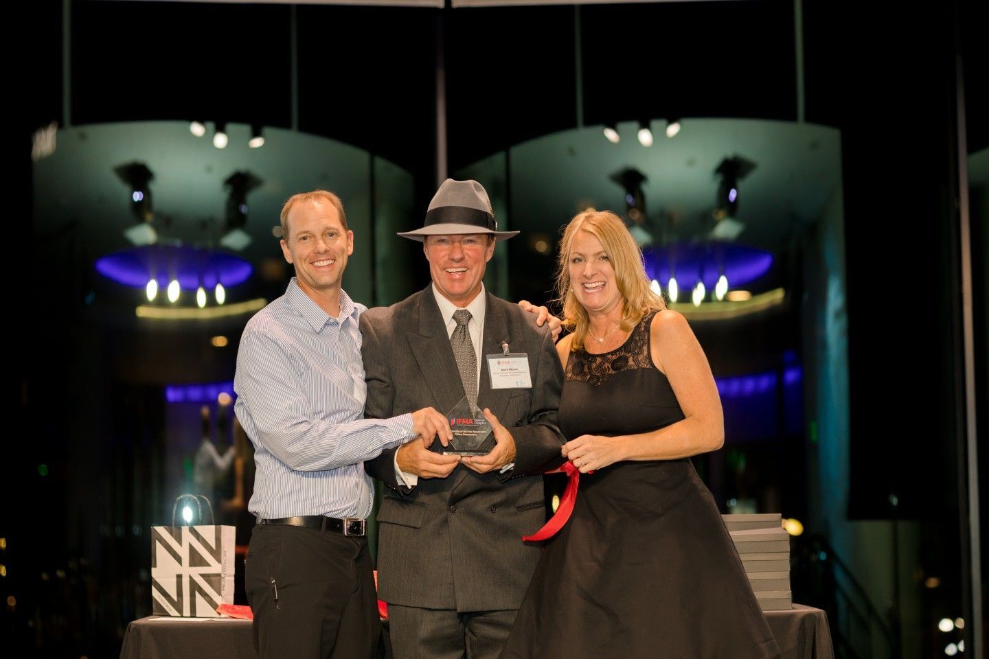 Three people on stage, one holding an award, smiling. Woman in black dress, man in suit, another man in a button-down shirt.
