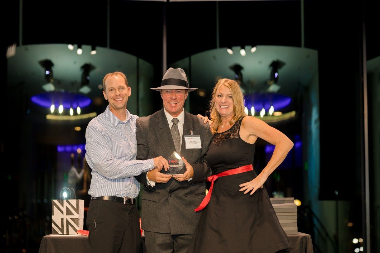 Three people on stage, one holding an award, smiling, and posing in front of a modern background with blue and white lights.