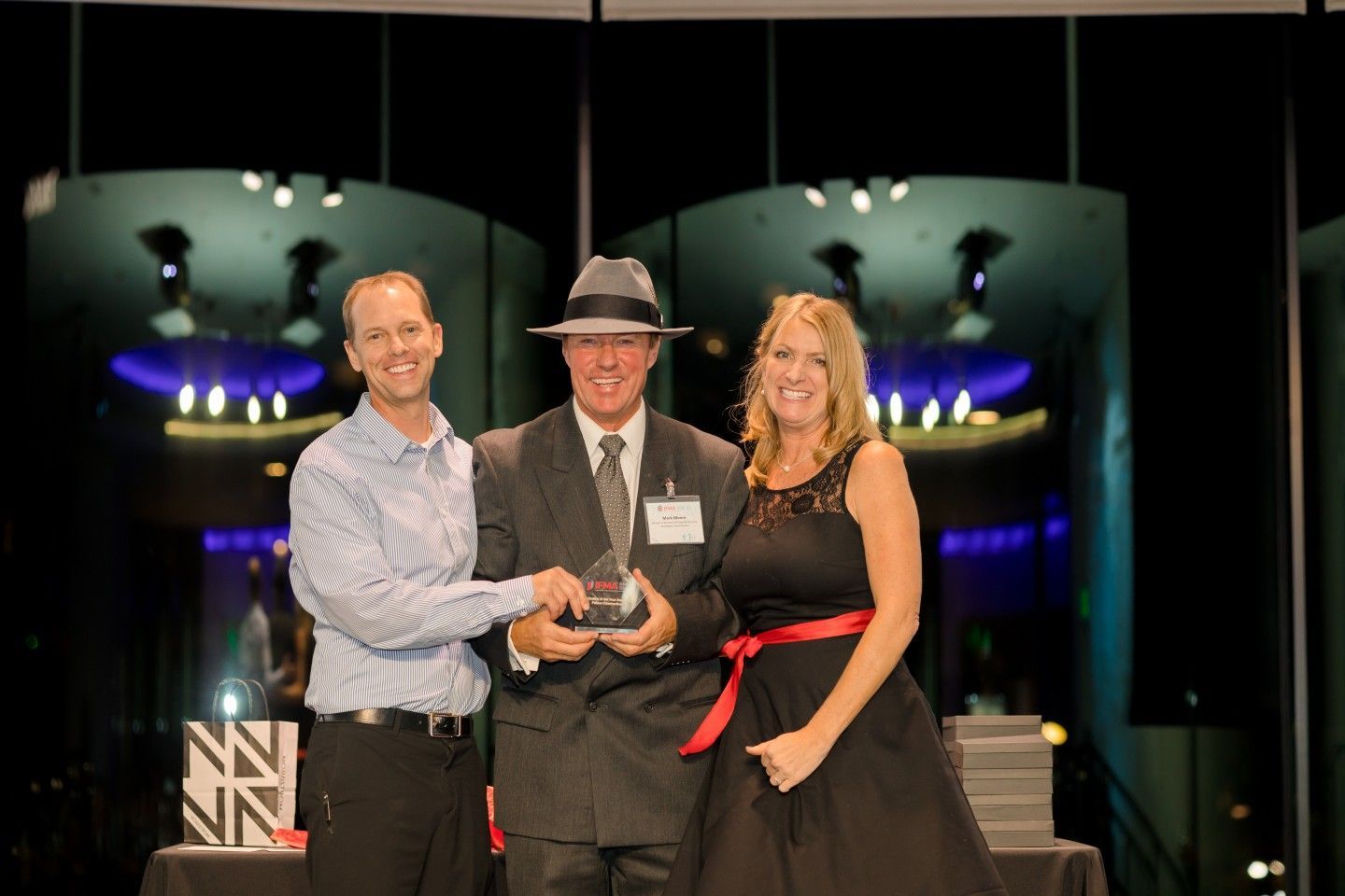 Three people on stage, one holding an award, smiling. Nighttime setting with colorful lights in the background.