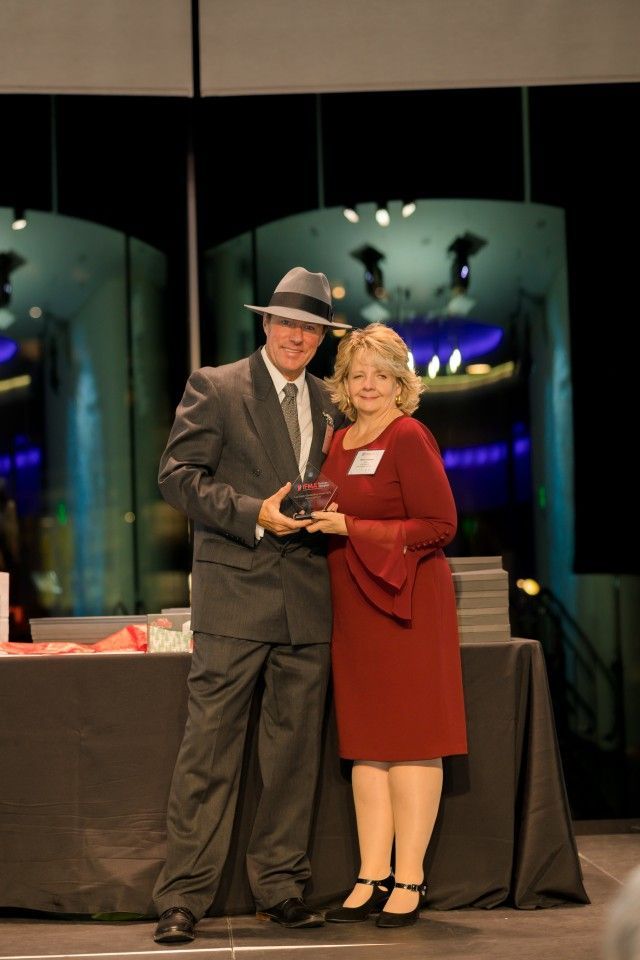 Man in suit and fedora presents award to woman in red dress.
