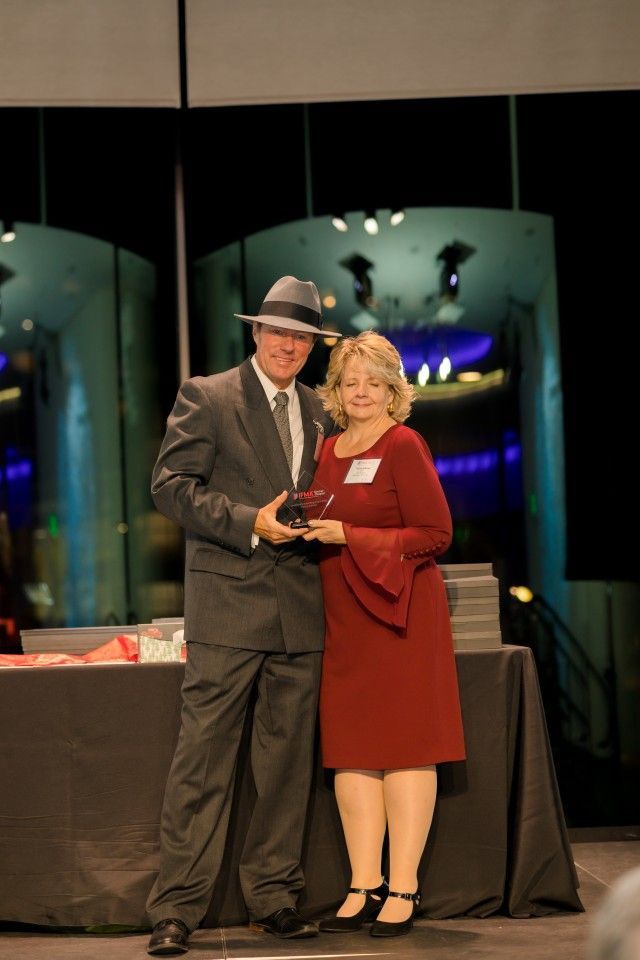 Man in fedora and suit presents award to woman in red dress.