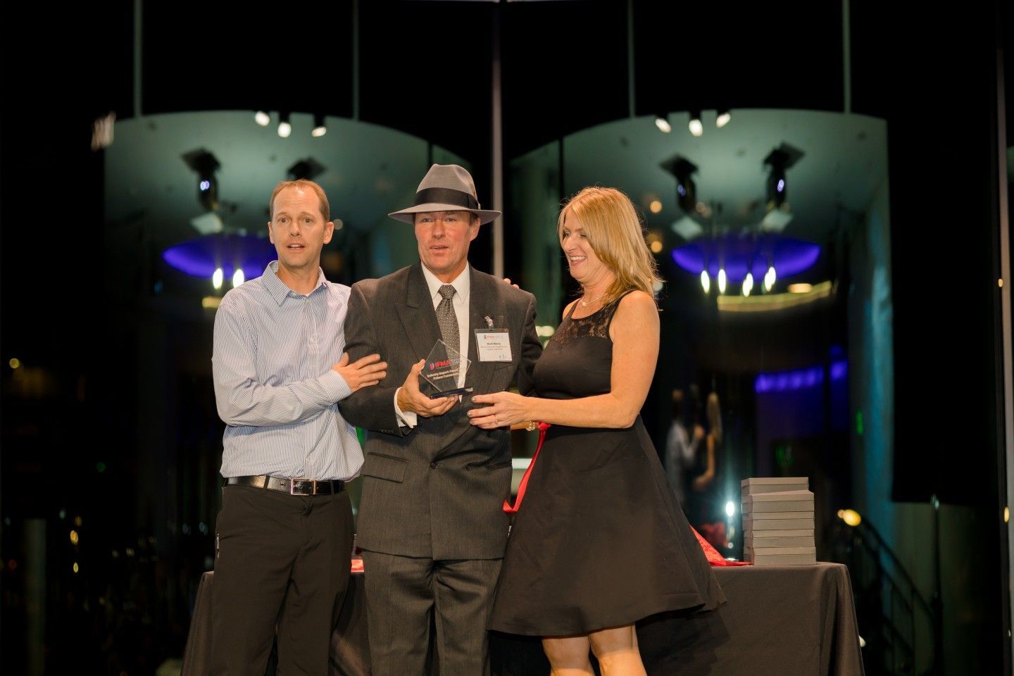 Three people on stage; man in fedora holding award, flanked by two others, at an event.