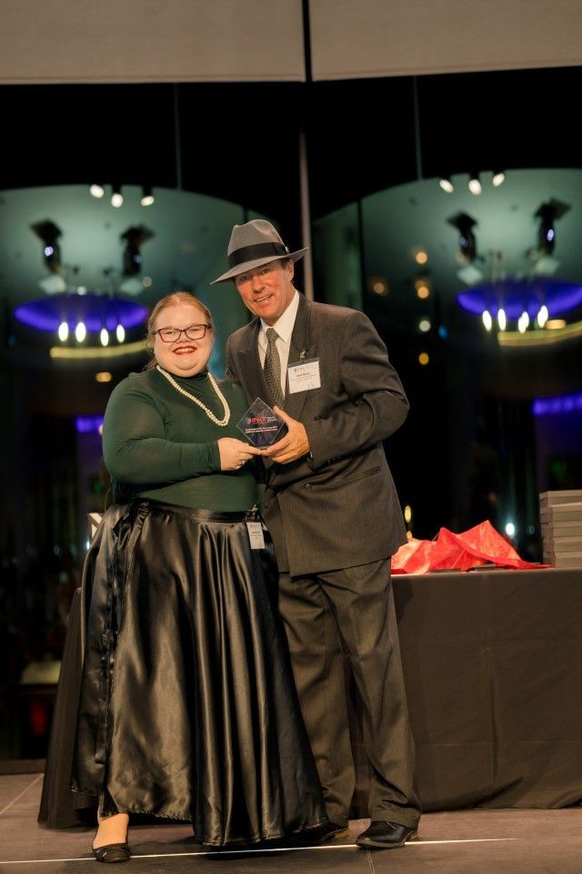Woman in long skirt and man in suit, both wearing hats, holding an award on a stage.