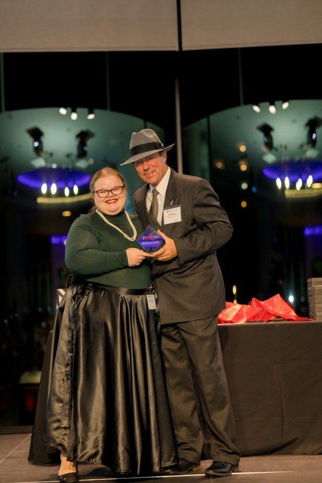 Person in green shirt and long black skirt accepts an award from a man in a suit and hat on stage.