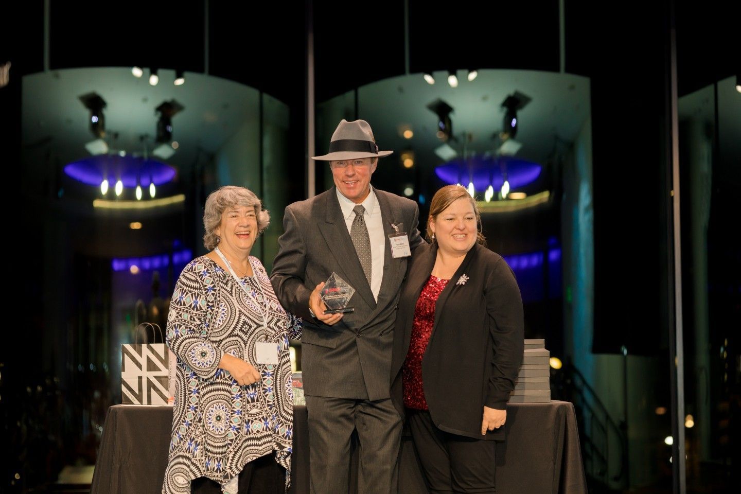 Three people on stage; man in suit holds award, flanked by two women, event setting.