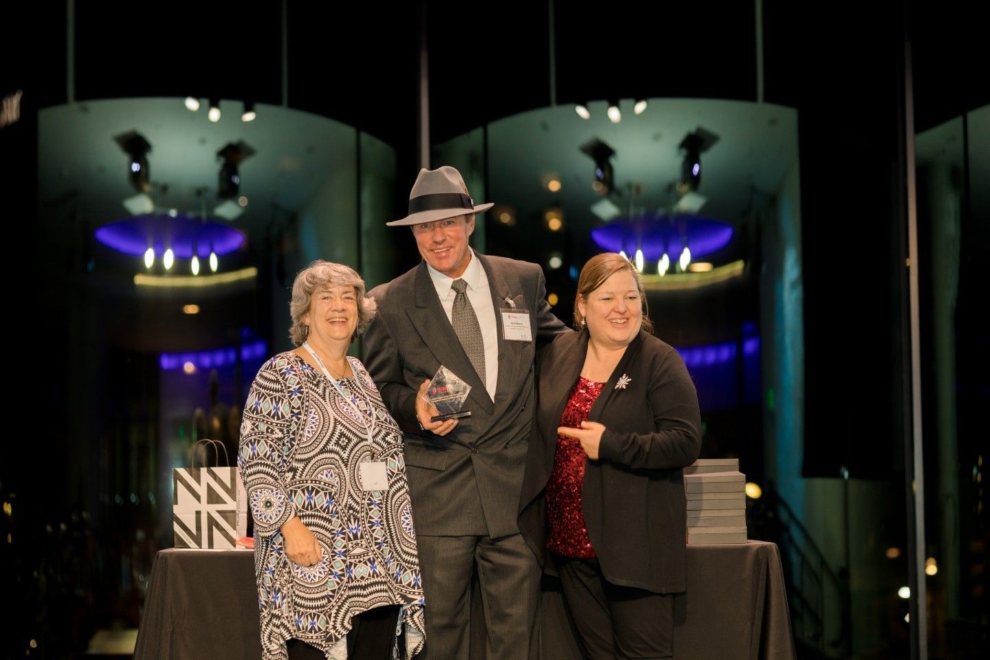 Three people on stage, holding an award. Man in suit and hat, flanked by two women, smiling, in front of stage backdrop.