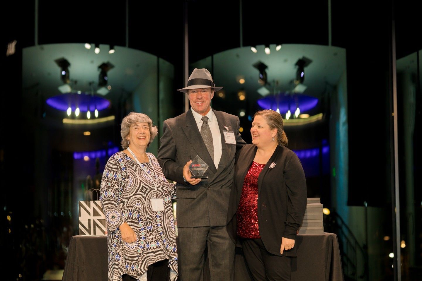 Three people on a stage; man in suit and fedora holds award; two women flank him.