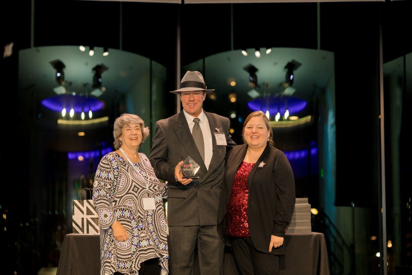 Three people stand onstage holding an award. Man in suit and fedora holds trophy, flanked by two women.