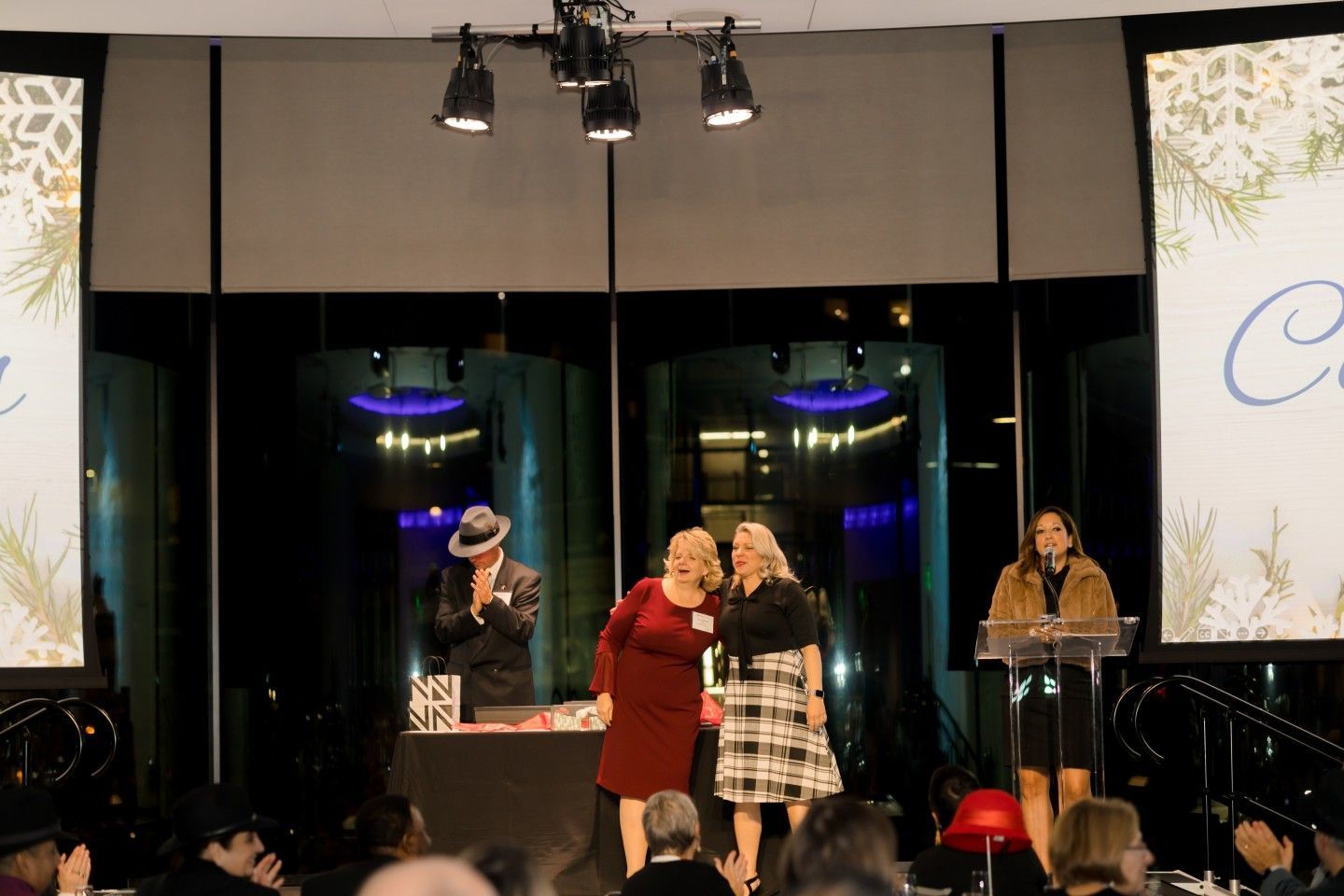 People on stage at an event: a magician, two women embracing, another woman at a podium. Black backdrop, stage lights.