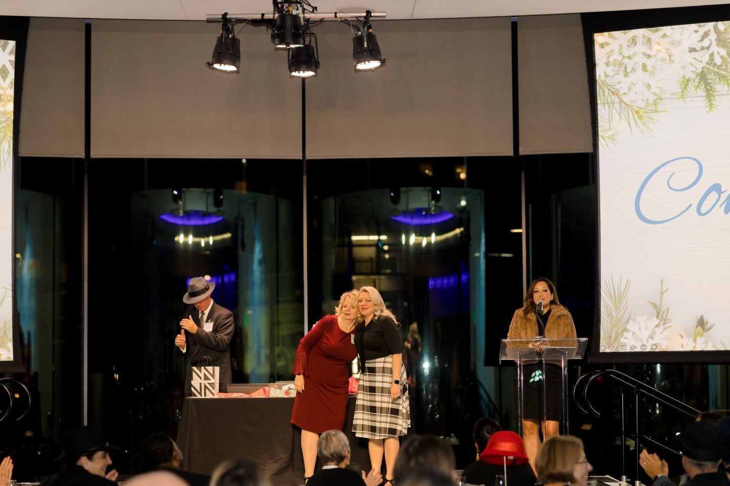 Two women embrace on stage at an event; a person speaks at a podium; a man stands nearby, behind a table.