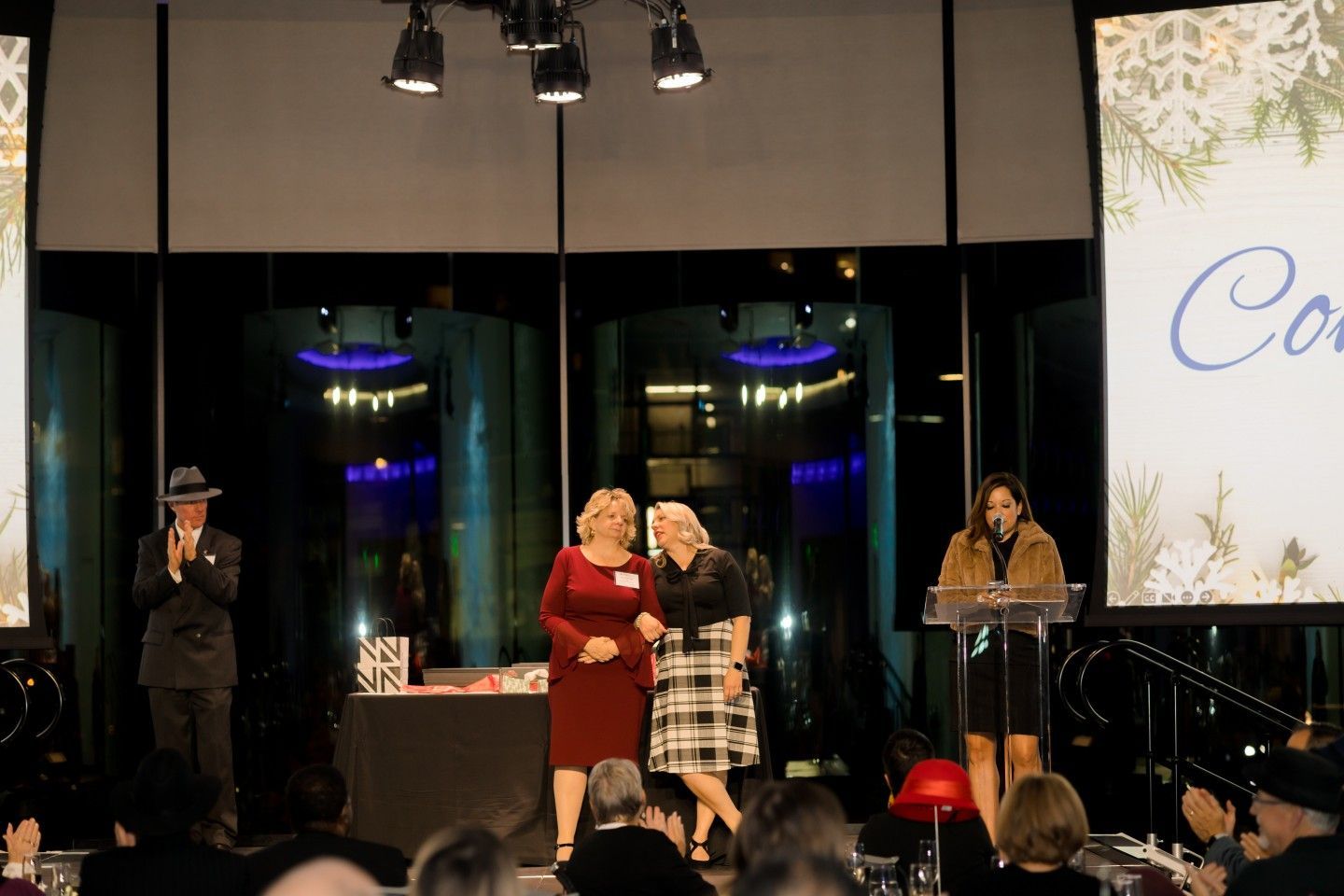 People on stage at an event: two women, a speaker, and a man in a hat. A screen shows 