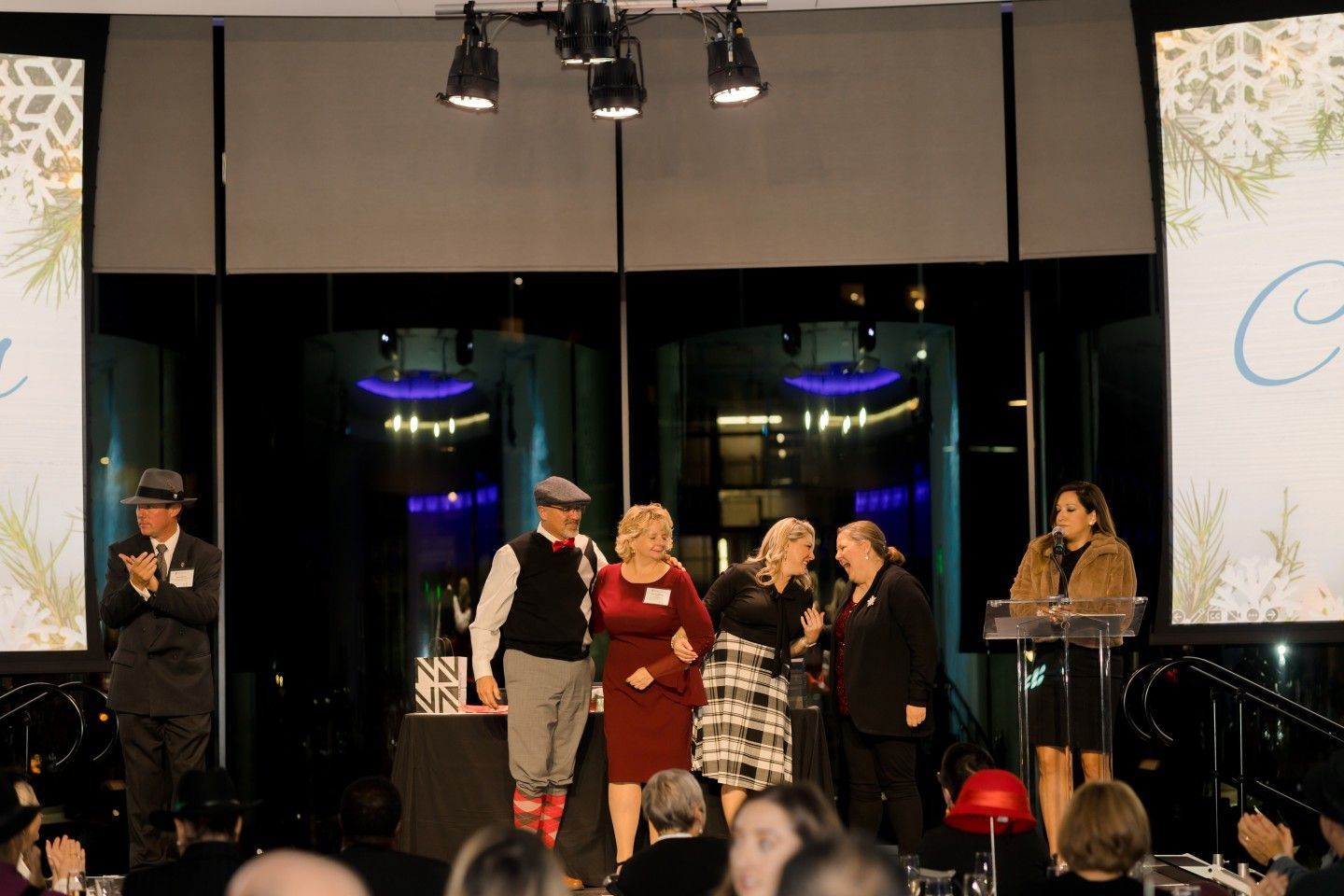 People on stage at an awards ceremony; woman speaking, others in festive attire, lit backdrop.