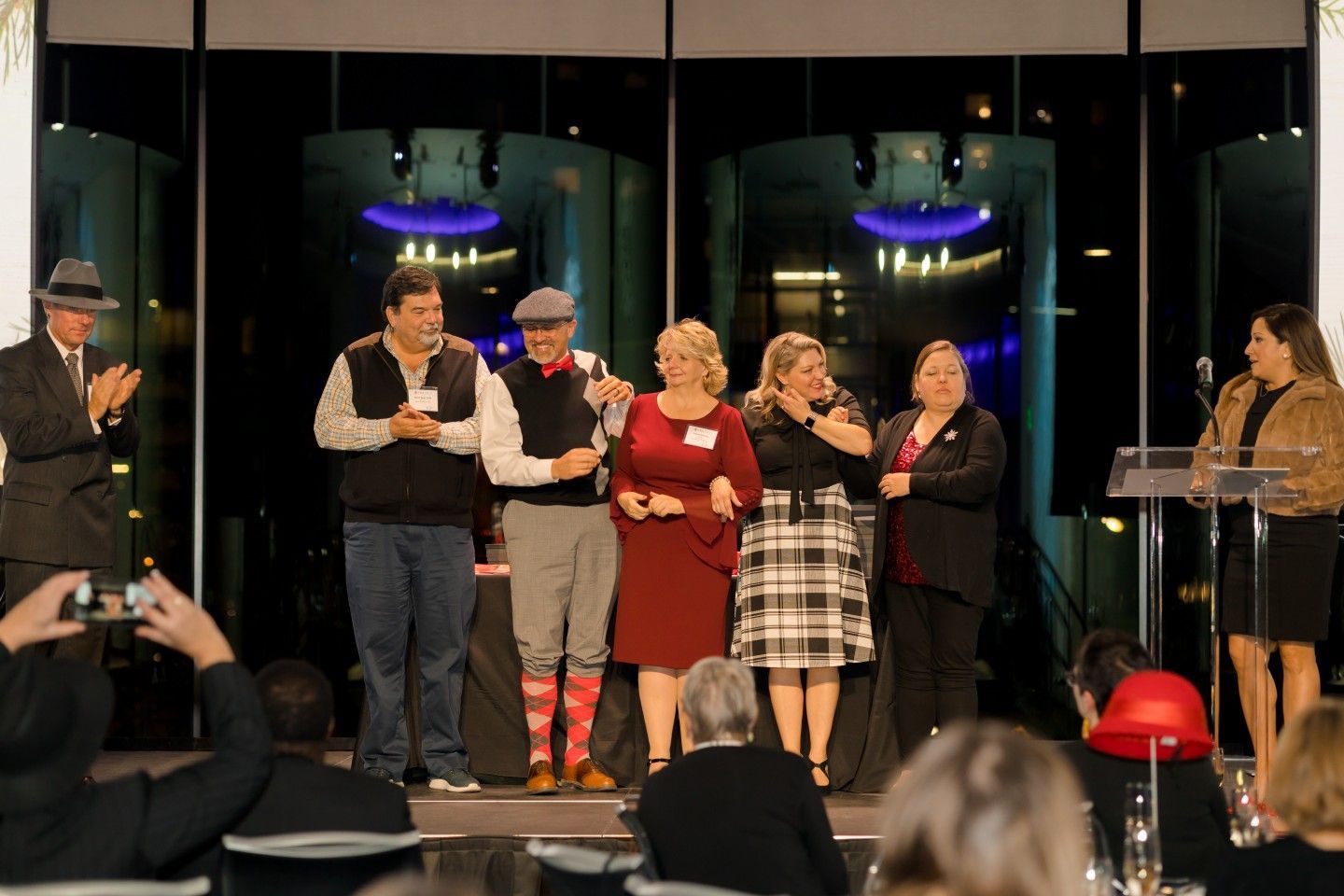 Group of people on stage at an event, clapping. Building with blue lights visible through windows.