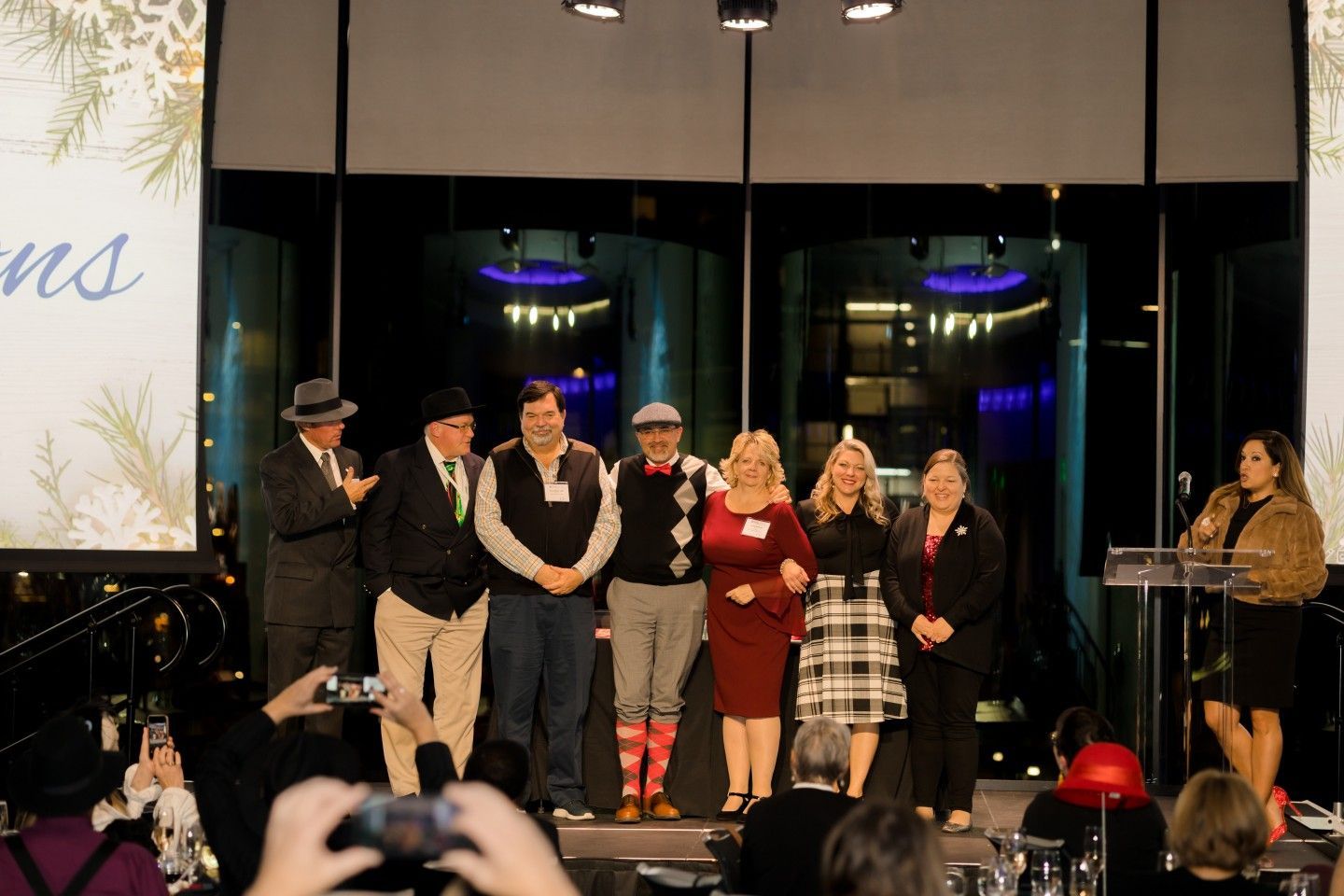 Group of people on stage, some with hats, posing for a photo. A woman speaks at a podium. Festive setting.